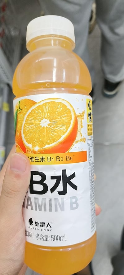 一只手握着一瓶外星人品牌的B水维生素饮料，饮料为香橙口味，瓶身呈橙黄色。标签上印有切开的橙子图案，突出显示“维生素 B1 B3 B6”字样，以及“B水 VITAMIN B”和品牌名“外星人 ALIEN ENERGY”。净含量为500mL。背景可见超市货架和模糊的行人腿部，推测拍摄场景为超市内。