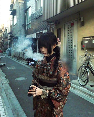 在充满日式市井氛围的东京小巷中，一位身着带有复古花纹和服（搭配毛绒腰封）的少女站在路中央，双手做出持握物品的姿势，身前飘出白色蒸汽，营造出类似吸食电子烟的现代行为场景。少女梳着带有粉色发饰的编发，面容清冷，与传统和服形成强烈的现代反差感。背景是典型的日式低矮民居、停在路边的自行车，远处还有休憩的行人，还原了东京小巷的日常市井气息。
注意：人物手部存在AI生成的错误（多根手指），这是AI生成图像的典型特征。