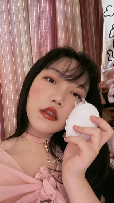 图中是一位长发女性的自拍，她手持白色美妆蛋靠近脸颊，妆容精致，唇色为橘红色，脸颊带有自然红晕。上身穿着粉色带蝴蝶结的上衣，脖颈佩戴粉色choker项圈，项圈上有金色链条装饰。背景是粉色条纹窗帘，右侧可见一个带有卡通云朵图案的白色袋子和一个粉色玩偶。整体画面风格柔和甜美，充满少女感，可能用于美妆或日常穿搭分享。