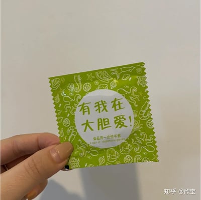 图片展示了一只手拿着独立包装的食品用一次性手套，包装为亮眼的绿色底色，印满白色的海鲜类简笔画图案，包括海马、章鱼、虾、贝壳、海星等，契合手套的使用场景。包装中央的白色圆形区域印着极具暧昧感的标语“有我在 大胆爱！”，下方标注了产品真实信息“食品用一次性手套”及英文“A pair of disposable gloves”。该包装模仿了安全套的包装风格与话术，用反差感的文案制造趣味，让普通的一次性手套变得诙谐有趣，打消人们在吃海鲜、小龙虾等食物时弄脏手的顾虑，同时带来轻松的体验。