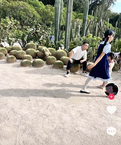 这是一张短视频平台的截图，拍摄场景为沙生植物园区，地面为沙地，背景分布着大量带尖刺的球状金琥仙人掌、高大的柱形仙人掌以及其他绿植。画面中一名穿白色T恤黑色长裤的男子误将带刺的金琥仙人掌当作可落座的物体，直接坐在上面，神情看起来十分错愕，疑似被刺扎到感到不适，旁边有两名小女孩正从此处路过。画面右侧带有短视频平台的互动UI，显示该内容获18.5万点赞、7619条评论，搭配配文“不是 大哥 你”，表达了对男子这一莽撞迷惑行为的诧异，整体充满喜剧效果。
