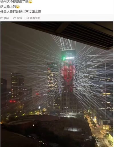 这是一张社交媒体截图，上方是网友的吐槽配文：“杭州这个楼是疯了吗😳 这大晚上的😳 外星人攻打地球也不过如此啊”，下方实拍了杭州夜晚的CBD城市景观，画面中心的中国人寿大楼被从楼顶发散出的大量白色激光线交织覆盖，密密麻麻的光束在夜空中形成复杂的网络，视觉效果极具冲击力，像极了科幻作品中外星人降临的场景，配文用夸张的调侃来形容这一奇特的灯光秀场面。