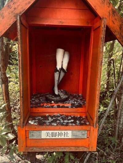 这张图片展示了一个独特的小型木制神社，通体呈鲜艳的橙红色，位于户外绿树环绕的环境中。神社的门是敞开的，可以看到内部供奉着一双穿着黑色渔网袜和高跟鞋的女性腿部雕像。神社底部的架子上和地面上散落着许多硬币，似乎是参拜者投掷的供品。神社正面有一块木牌，上面写着“美脚神社”四个汉字，表明这是一个专门供奉“美脚”的独特神社。整个场景给人一种奇特而有趣的感觉，融合了传统神社形式与现代审美元素。