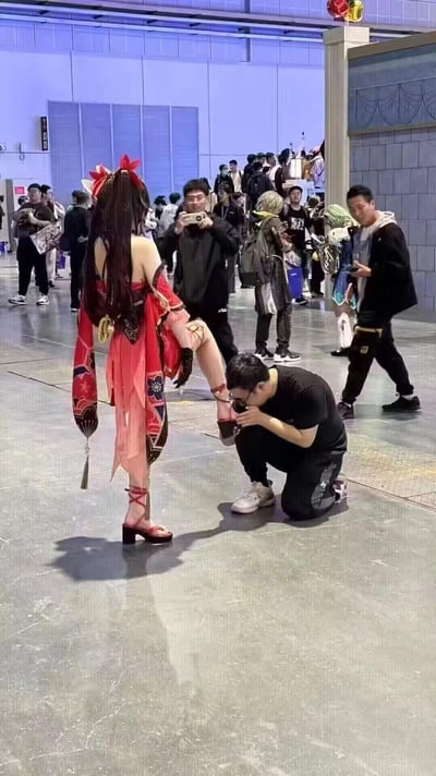 这是在一场国内大型漫展的现场，一位女性coser还原了《崩坏：星穹铁道》中的角色花火，她单腿抬起站立，一名男性粉丝跪在地上，做出亲吻她鞋子的举动。周围聚集了不少漫展参与者，有人在拍照记录，有人在围观，现场人来人往，氛围热闹，这个举动也引发了不少争议。