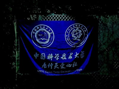 这是一张户外实拍的中国科学技术大学唐仲英爱心社旗帜照片，背景为铁丝网围栏。旗帜主体为深蓝色，上方左侧是带有1953建校年份的中国科学技术大学校徽，右侧是唐仲英爱心社的专属社徽；下方依次印有“中国科学技术大学”“唐仲英爱心社”的中文名称，以及对应的英文“USTC Cyrus Tang CaringHeart Club”，整体风格正式庄重，体现了社团的公益属性与校园归属感。
