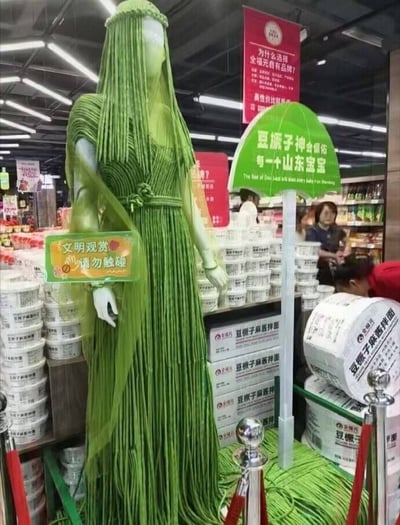 这张实拍图拍摄于山东全福元超市内部的食品售卖区，是超市结合本地特色打造的趣味营销场景：核心展示物是用大量绿色长条形豆橛子（长豆角）装饰的人形模特，豆橛子模拟出神像的长发与长袍效果，整体为通体绿色的拟人化神像造型。模特旁立有两块提示牌，一块提示“文明观赏 请勿触碰”，另一块伞形绿色提示牌写有玩梗文字“豆橛子神会保佑每一个山东宝宝”，造型周边用隔离栏围挡，周边货架上堆叠摆放着全福元品牌的豆橛子麻酱拌面商品，背景可见明亮的超市照明、其他宣传标语以及正在购物的消费者。