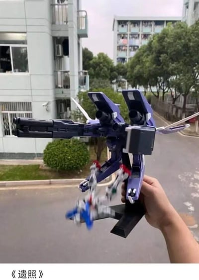 图中展示了一只手握着一个大型紫色高达模型，模型装备着巨大的武器系统。在模型前方，一个小型蓝白相间的高达模型呈现出被击中后坠落的姿态，动作十分生动。背景是校园环境，有教学楼、绿化带和道路。图片底部配有文字《遗照》，形成幽默效果。

双关暗示：标题《遗照》与模型坠落的姿态形成搞笑对比，将玩具模型场景夸张地比作战场遗照。