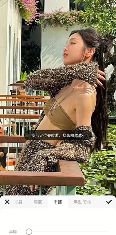 这是一张手机修图软件的操作截图，画面主体是一位身处户外露台（配有水景、绿植与休闲设施）的女生，她身着卡其色比基尼外搭混色针织衫，姿态为单手搭在颈后，另一只手置于栏杆上，神情放松。软件底部栏显示「瘦身」「肩颈」「丰胸」「手动美体」等美体修图选项，界面中间弹出提示文字：「胸部定位失败啦，换张图试试~」。由于女生的手臂遮挡和身体角度，导致修图软件的AI无法识别胸部位置，出现功能失效的搞笑场景。