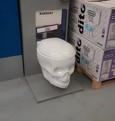 图片中展示了一款造型独特的白色马桶，其外观被设计成人类骷髅头的形状，顶部为光滑的圆形“头骨”，正面清晰可见骷髅面部轮廓及牙齿细节。马桶安装在灰色的方形底座上，后方连接着带有“GEBERIT”品牌标识的白色水箱，水箱上贴有红色标签。右侧堆放着多个印有“dito 2”和“Rimfree”字样的产品包装盒，盒子上印有马桶的示意图及相关认证标志。整体场景疑似家居建材店或产品展示区，突出展示了这款极具创意和视觉冲击力的骷髅头造型马桶。