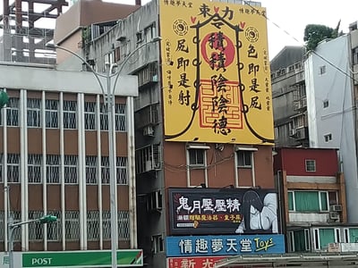 这是拍摄于台湾地区街头的户外广告场景，一栋老旧居民楼外悬挂着多层情趣用品店「情趣夢天堂」的广告牌。最上方的黄色广告牌模仿道教符咒的视觉样式，玩起低俗谐音梗：将「积阴德」改为带有暗示性的「積積陰陰德」，同时把佛教名句「色即是空，空即是色」恶搞成「色即是屁，屁即是射」，搭配侧边的戏谑小字，用传统文化的包装形式做成人用品的擦边宣传；下方的电子屏则结合台湾鬼月的民俗，打出「鬼月壓壓精」的促销标语，声称满额赠送「驅魔法器、童子尿」，延续恶搞营销的风格。整体广告通过传统文化与成人用品宣传的强烈反差，加上低俗谐音的戏谑感，极具魔性，吸引路人注意力。
谐音梗说明：「積積陰陰德」谐音「积阴德」，「積積」暗指男性生殖器官；「色即是屁」「屁即是射」谐音「色即是空，空即是色」，玩低俗擦边球。