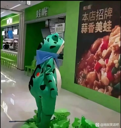 这是一张商场内的实拍静态图，场景位于主打美蛙菜品的“蛙喔”餐厅门口。一个身着绿色带黑斑青蛙人偶服的角色站立在餐厅门前，抬头凝视着外墙上的“本店招牌 蒜香美蛙”宣传海报，海报上展示着烹制完成的美蛙肉。人偶的脚下还摆放着几个小型的青蛙玩偶，整个画面形成了极具荒诞感的反差：作为餐厅吉祥物的青蛙，正看着自己的同类被做成招牌菜品的宣传图，仿佛“打工人发现自己的工作是为出卖同类站台”，这种荒诞的错位感构成了地狱笑话式的笑点，戳中了职场自嘲的情绪，显得滑稽又讽刺。