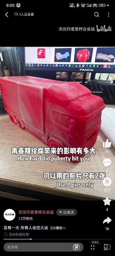这是一张哔哩哔哩（B站）的视频播放页截图，画面主体是放置在木质桌面上的红色半透明卡车造型硅胶类物品，背景可见熊猫品牌的电脑显示器，屏幕上展示了多款同类硅胶制品的商品图，桌面还摆放有键盘、笔记本等物品。画面叠加了两行中英双语玩梗文字：“青春期给你带来的影响有多大 How hard did puberty hit you”、“可以用的照片只有2张 Use 2 pics only”，利用英文原句中“hit（击打、撞击）”的含义，用卡车对应“撞击”的意象制造笑点。界面显示该视频的发布者为B站UP主“次元行者圣杯企业店”，视频标题为“总有一天 所有人会控大运”，标注有“含AI生成内容”，播放量为225次，账号粉丝量为1.2万，界面右侧带有B站常规的点赞、评论、投币、收藏、转发功能按钮，底部有弹幕发送栏。