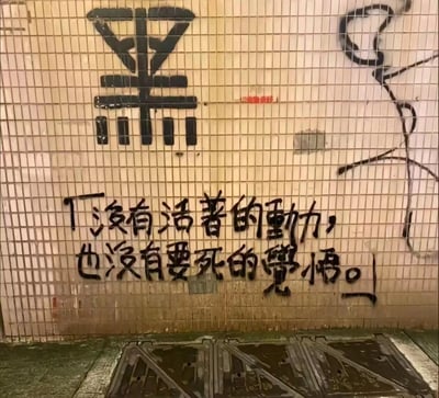 一张城市街头的摄影照片，展示了一面带有网格纹理的墙壁。墙上有黑色涂鸦，最上方是一个大型的"黑"字，下方则是一行手写风格的文字："没有活着的动力，也没有要死的觉悟。" 整幅作品呈现出一种都市年轻人中常见的迷茫与灰色情绪，反映了对生活状态的无奈与困惑。画面下方可以看到地面和排水井盖，暗示这可能是在地下通道或类似的城市空间拍摄。