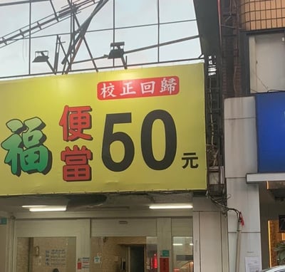 这是一张街头便当店门口的实拍图，画面主体是一块明黄色的店铺招牌：招牌顶部的红色矩形框内写着“校正回归”，下方左侧是绿色的“福”字，旁边是红色的“便当”字样，右侧是醒目的黑色大号字体“60元”，清晰传递出这家“福便当”店铺将便当价格回调至60元的信息，主打亲民平价的餐食。招牌下方是店铺的入口区域，能看到店内的灯光和部分墙面设施，整体呈现出台湾街头常见的平价餐饮小店的日常风貌，没有双关、谐音等特殊梗，是直白的价格调整宣传。