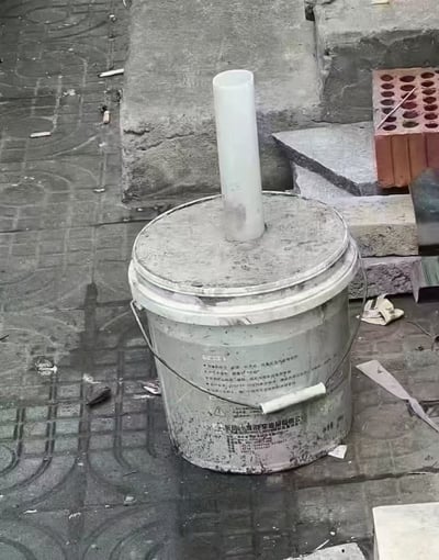 这是一张拍摄自工地或类似环境的图片，画面中央是一个带盖子的油漆桶，桶盖中心处插着一根白色塑料管。桶身有明显的使用痕迹和污渍，四周散落着建筑材料、砖块等杂物。整个场景给人一种简陋、随意但充满生活智慧的感觉。

图片中的桶和管子组合很容易让人产生联想：乍一看像某种科学实验装置，或暗示诺贝尔奖的‘新发明’，实际上则是常见的工地简易烟灰缸。这种“质朴但别有用心”的设计，带有一定的幽默感和生活双关意味。

暗示：画面通过“看起来很科学”的装置表达对生活智慧的幽默讽刺，有调侃‘工地诺奖’、“发明家精神”的双关含义。