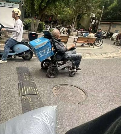 街头场景中，一位 elderly man 坐在代步车上，车后装有蓝色的美团外卖箱，正在道路上行驶。其左侧有一名戴着黄色头盔、身穿白色上衣的男子骑着蓝色电动车同行。背景可见树木、长椅，有行人休息，路边停放着多辆电动车，远处有带有中文标识的招牌，整体呈现日常生活气息浓厚的街景。