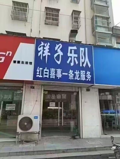 这是一张线下商铺的实景拍摄图，画面核心是位于居民楼底层的一家商铺，其蓝底招牌上用白色大字写着“祥子乐队”，下方搭配红色小字“红白喜事一条龙服务”，表明该店铺承接婚礼、葬礼相关的乐队服务。左侧相邻的是带有“智慧生活馆”标识的商铺，“祥子乐队”店铺的玻璃门内还能看到“新总窗帘”的字样，门口外墙上挂着一台白色空调外机，画面右下角露出了一辆电动车的局部，背景是带有防盗窗的居民楼，整体是日常的城市街道商铺场景，没有双关、谐音梗等特殊内容。