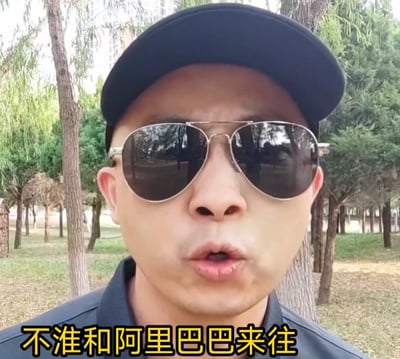 户外场景中，一名戴着黑色鸭舌帽和墨镜的男子站在树木环绕的草地上，背景可见多棵绿树和远处的建筑物。男子面部表情严肃，嘴巴微张呈说话状态，图片底部有醒目的黄色文字'不准和阿里巴巴来往'，整体画面给人一种命令式的严肃氛围。