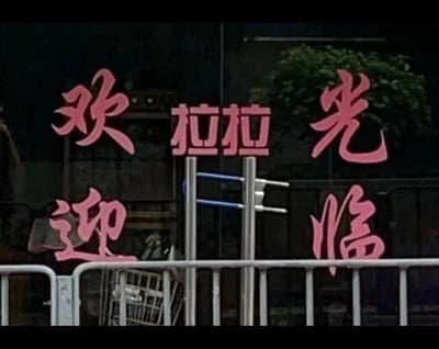 夜晚拍摄的店铺招牌照片，粉色霓虹灯字体显示'欢拉拉光'和'欢迎临'字样，背景可见玻璃橱窗和绿色植物。