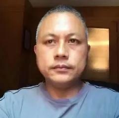 这是一张室内拍摄的中年男性正面肖像照片，男子头发花白，穿着浅灰色带深色领口的上衣，面部表情严肃，双眼直视镜头，背景为室内的木质墙面与门，整体构图清晰，画面焦点集中在人物面部。