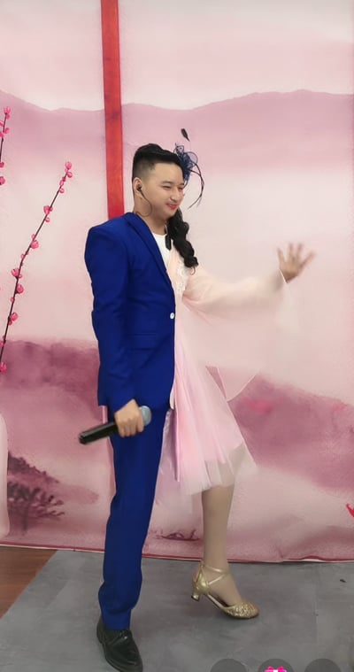 这是一段创意表演的画面，表演者通过特殊的服装造型实现一人分饰男女两角的视觉效果：身体左侧是身着宝蓝色西装、佩戴耳返的男士造型，右手持麦克风；右侧是身着粉色古风裙装、头戴深色发饰的女士造型，左手抬起做出挥手动作。表演者面带微笑，神态愉悦，背景是带有粉色梅花枝的古风布景，整体利用服装和姿态制造出一人同时呈现两个角色的趣味错觉，场景大概率是短视频平台的直播表演，风格诙谐有趣。