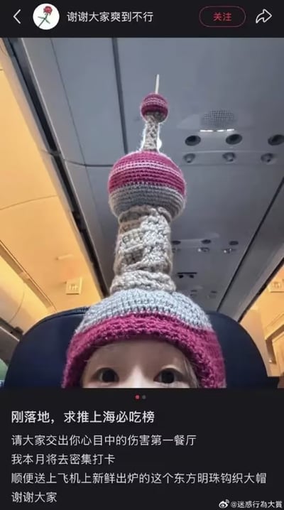飞机机舱内，一人头顶戴着造型夸张的东方明珠塔钩织帽——塔身由粉、灰、白毛线层层编织，顶部尖刺还原度极高，帽檐为粉色。配文称刚落地上海，求推荐必吃餐厅（戏称“伤害第一餐厅”），并调侃这顶飞机上“新鲜出炉”的手工帽。