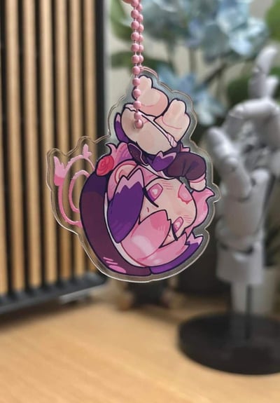 这是一个亚克力材质的Q版角色挂饰，角色以粉紫为主色调，蜷缩成一团，双眼为爱心样式，头顶带有粉色光环装饰，胸口还有小玫瑰点缀，被粉色珠链悬挂着。背景是室内桌面场景，能看到模糊的手办和装饰物件，整体风格软萌可爱，充满二次元气息。