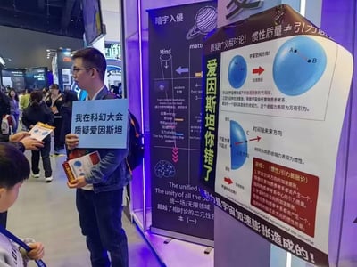 这是在国内科幻大会现场的实拍场景：一名戴眼镜的男子手持写有「我在科幻大会质疑爱因斯坦」的蓝色标语牌，同时拿着印有相关理论的宣传册，站在一块写有「爱因斯坦你错了」的展板旁，展板上以《惯性/引力膨胀论》为核心，质疑爱因斯坦的广义相对论，将万有引力解释为宇宙空间膨胀的收缩力，惯性解释为时间方向的收缩力，试图构建新的物理理论体系。现场还有其他参会人员穿梭，整体是公开展会中个人展示学术质疑观点的场景。
