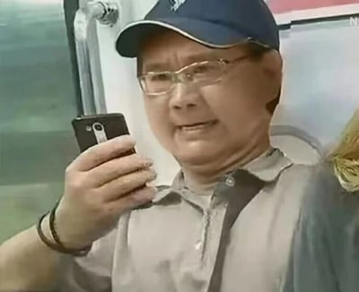 图片中一位戴着蓝色鸭舌帽和眼镜的中老年男性，身穿浅灰色衬衫，正用右手拿着一部黑色老式智能手机，似乎在查看屏幕或准备拍照。背景模糊，隐约可见类似交通工具内的座椅和窗户，整体氛围日常随意。