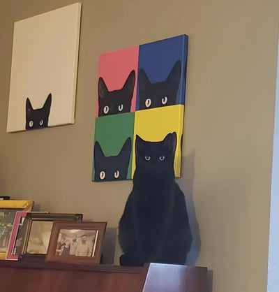 室内场景中，一只纯黑色猫咪静坐于木质书架上，眼神专注凝视前方。猫咪正上方的米色墙壁上悬挂着两幅猫咪主题油画：左侧为白色画布上的黑色猫咪头像剪影（仅露出耳朵和圆睁的双眼），右侧则是由粉色、蓝色、绿色、黄色四色方块组成的拼贴画，每个色块中均有一个姿态相似的黑色猫咪头像。现实猫咪与画作中的猫咪形象在姿态和眼神上形成奇妙呼应，营造出幽默又温馨的视觉效果。画面下方书架上还摆放着多个相框，增添了生活气息。