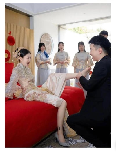 这是一张中式婚礼现场的照片，画面中新娘身穿传统秀禾服坐在婚床上，单腿抬起，身穿高跟鞋，丝袜上写有一个明显的“S”字母。新郎单膝跪地，正要为新娘穿鞋。背景有四位伴娘身穿统一服饰站立，房间装饰有明显的中国风元素如红色悬挂饰品和红床单。地上散落一些纸币，可能象征“接亲”环节的讨红包或仪式。照片氛围喜庆且稍带趣味感。

亮点：新娘丝袜上“S”字母成为全图最具梗感的细节，很可能具有关联词“丝袜”/“S号新娘”或“Super”等双关，用于调侃或玩笑。

【谐音/双关梗】：“S”暗示丝袜、尺寸也可理解为“Super”，在婚礼梗图中颇有话题度。