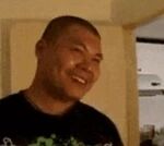 图片中是一位光头男子，穿着深色上衣，正面向镜头露出微笑。背景为室内环境，可见米色墙壁和一些模糊的背景元素，整体氛围较为日常亲切。