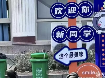 这是一张校园迎新场景的实拍静态图，画面主体是一根橙色立柱，立柱上有两组蓝底白字的圆形标语，分别是“欢迎你”和“新同学”，用于迎接新生。立柱下方有一个指向左侧的箭头标识，标识上写着“这个最美味”，而箭头恰好指向旁边的绿色厨余垃圾桶，形成了极具趣味的错位效果，仿佛在调侃垃圾桶里的东西是“最美味”的，制造出搞笑的乌龙感。背景中可见学校建筑物的外墙、空调外机，还有一些绿植，旁边还有另一个绿色垃圾桶。画面右侧有一个面带微笑、脸颊泛红的卡通宇航员形象装饰，右下角带有安徽工业大学的相关水印，整体场景充满校园生活的趣味感。