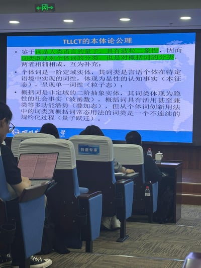 学术会议现场，参会者坐在标有“特邀专家”的座位上，部分人使用笔记本电脑。前方大屏幕显示“TLLCT的本体论公理”，内容将词比作“人类语言的量子”，提出其具有波粒二象性，论述个体词（一阶定域实体，粒子态）与概括词（二阶抽象实体，叠加态）的词类分类及相辅相成关系，涉及认知事实、社会事实、量子跃迁等概念，部分文本用绿色高亮。