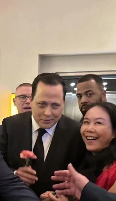 这是一张因视觉错位产生趣味效果的合影照片。画面中，一位身着黑色西装、别着粉色康乃馨的男士站在中间，他身体后方两侧的两位男士头部，因位置巧合恰好和中间男士的肩膀重合，营造出中间男士拥有三个头的错觉。旁边的女士笑容灿烂，看起来对这个有趣的错位效果感到开心。场景为室内正式活动现场，推测可能是婚礼或晚宴场合，整体氛围轻松欢乐。