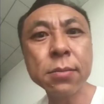 这是一张近距离拍摄的男性脸部特写静态图，图中人物是中文互联网知名梗人物神鹰哥（神鹰黑手哥），他眉头微皱，正视镜头，表情严肃，穿着白色上衣，背景是浅色带斜纹分格的瓷砖墙面，这是神鹰哥流传最广的经典原始大头素材图。