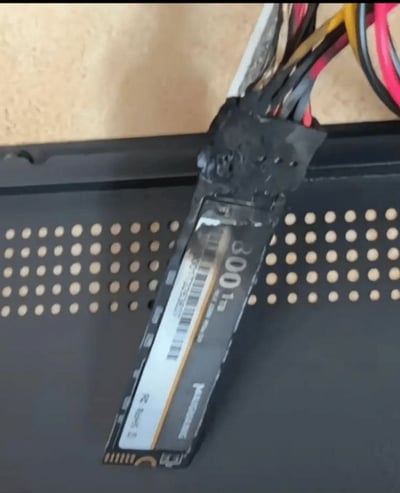 一张展示被烧毁的Panasonic SSD固态硬盘的照片。SSD的顶部区域严重烧焦变黑，外壳有明显的灼烧痕迹，部分线路从烧焦处引出。硬盘下方是带有均匀圆孔的黑色金属面板，背景隐约可见木质桌面。整体画面直观呈现了电子元件因故障或短路导致烧毁的状态。