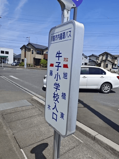 日本埼玉县川越市的一处街道实拍，画面中央竖立着一块市内循环巴士指示牌。牌面顶部标注“川越市内循環バス”，主体部分以蓝色文字写有“牛子小学校入口”，并配有红色箭头指示方向，下方辅以“次は旭橋東”（下一站旭桥东）和“藤木”等地名信息。背景可见日式传统住宅建筑群、停放的白色轿车及晴朗蓝天下的电线，展现典型的日本郊区日常街景。