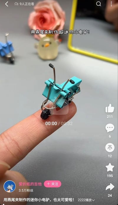 这是抖音平台的短视频截图，展示了一个用燕尾夹手工制作的超迷你小电驴，它小巧到可以稳稳站在人的指尖上，造型还原了电动车的结构，十分可爱。画面背景中还能看到同款小手工和小鸡造型的小摆件，视频创作者为“爱折纸的吉他”，视频时长32秒，是介绍该创意手工的制作内容，整体风格治愈有趣，适合手工爱好者观看，目前这条视频已有2222次播放，收获了不少点赞和收藏。