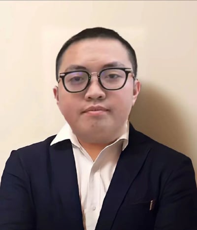 图片中是一位戴着黑色框架眼镜的男子，他穿着深色西装外套和白色衬衫，头发较短，背景是浅色的墙壁。男子正面朝向镜头，表情看起来比较平静自然。