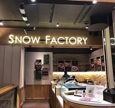 这是一张SNOW FACTORY店铺的内部环境照片。照片中可见店铺入口上方有白色发光的'SNOW FACTORY'招牌，店内采用木质装修风格，右侧为弧形点餐柜台，柜台上摆放收银机、菜单及纸巾等物品。柜台后方墙壁有石质装饰，挂有多幅食品展示菜单图片。左侧墙壁有绿色植物装饰和小架子，店内灯光明亮，氛围温馨，柜台后有工作人员正在忙碌。