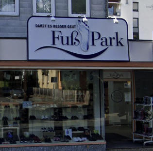 这是一张实拍的街景照片，展示了一家德语区的FußPark鞋店外观。店铺招牌采用蓝白配色设计，上方印有德语标语“DAMIT ES BESSER GEHT”（意为“让一切变得更好”），中间搭配脚印图案与店名“FußPark”——“Fuß”在德语中意为“脚”，直观点明店铺主营鞋类商品的属性。玻璃橱窗内整齐陈列着各类鞋款，橱窗右侧的外置鞋架上也摆放着鞋子，背景是浅灰色的多层建筑外墙，整体画面构图清晰，能清晰看到店铺的经营信息与陈列的商品。