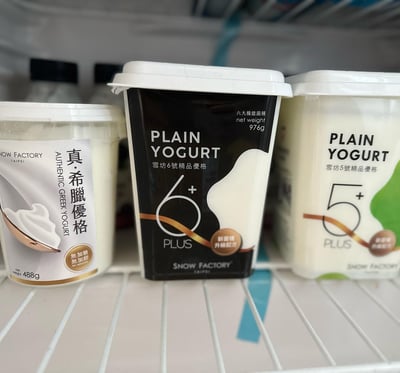 冰箱冷藏层的白色搁架上并排放置着三款雪坊(SNOW FACTORY)品牌的酸奶产品。左侧为白色包装的“真·希腊优格”，标注“无加糖”，净含量488g，包装上有酸奶勺的图案；中间是黑色包装的“雪坊6号精品优格”，主打“6+ PLUS”和“六大机能菌”，净含量976g，标注“新菌種升級配方”；右侧为白色带绿色点缀的“雪坊5号精品优格”，标注“5+ PLUS”。三款产品均为白色盖子，包装设计简洁，主要展示产品名称和核心卖点。