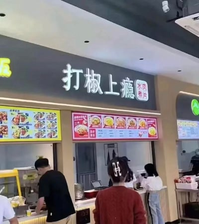 这是一张商场美食城档口的实拍图，档口名为“打椒上瘾”，主打水煮肉片。画面中一位穿红色上衣的顾客背对镜头，她的发夹是反向的“68”，视觉上看起来像是“89”，和档口名称的“打椒”形成谐音巧合，仿佛在呼应“打椒（89）上瘾”，制造出趣味效果。档口前还有其他顾客在点餐，上方的电子菜单展示了各类餐品，整体场景是热闹的美食城环境。
趣味点说明：反向发夹“68”视觉上近似“89”，与“打椒”谐音，形成偶然的趣味梗。