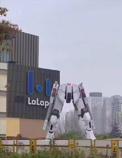 这是一张在上海LaLaport金桥店外拍摄的视觉错位照片，商场外立面上的蓝色品牌标识，与下方的实物大自由高达立像的下半身恰好通过拍摄角度重合，看起来仿佛商场的logo变成了高达的上半身，形成了极具趣味的视觉错觉，背景是城市居民楼，整体画面充满搞笑感，成为高达爱好者和打卡爱好者喜爱的趣味内容。