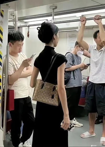 这张图片拍摄于地铁车厢内，画面中央是一位穿着黑色长裙的女性，手挎Coach包包，打扮精致、气质出众。周围乘客都较为休闲，或低头玩手机、或拉着扶手显得随意，形成鲜明对比。这种时尚与日常的反差让人眼前一亮，容易引发“地铁遇见时尚女神”等网络讨论话题。有明显的都市通勤生活气息和些许轻松幽默的都市反差感。整体氛围无奈中透露出一丝趣味。未见明显双关或谐音梗，但存在“日常地铁偶遇时尚模特级美女”的潜台词。