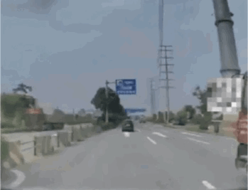 这是一张从车辆驾驶视角拍摄的公路画面，画面中是一条开阔的双向公路，前方有一辆黑色车辆正在行驶，道路左侧设有护栏和绿植，远处立有蓝色的交通指示牌，画面右侧存在被马赛克处理的区域，天空呈现灰蒙状态，整体呈现出日常公路出行的普通场景。