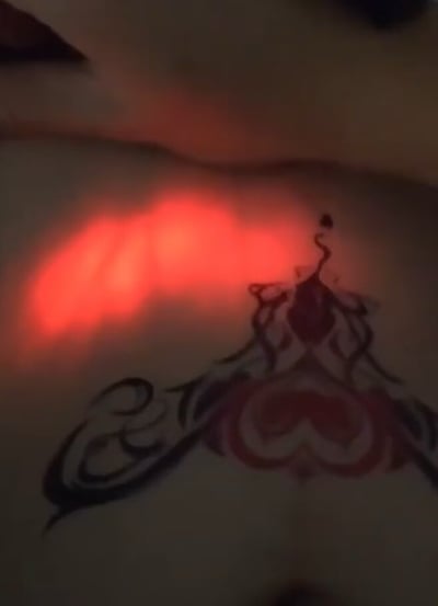 图片展示了身体某部位的纹身图案，纹身设计包含复杂的线条和红色心形元素，旁边有明显的红色光斑，疑似手电筒照射效果。标题暗示了某种不当行为，结合画面内容可能存在引人不适的暗示，需谨慎观看。