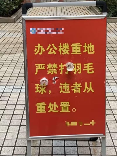 图片展示了一个放置在室外方格瓷砖地面上的红色可移动标语立牌，立牌带有金属边框，上面用黄色字体标注着“办公楼重地 严禁打羽毛球，违者从重处置。”的禁令内容。但极具反差感的是，标语牌上有两处被羽毛球击破的孔洞，还有两个羽毛球分别粘在标语的“打”字和“球”字附近，形成了被禁止的行为反过来“破坏”禁令标识的搞笑场景，充满了讽刺和趣味，像是对这条禁令的生动调侃。