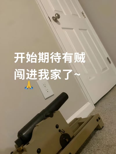 图片展示了一个室内场景，画面中央是一扇白色的门，门旁墙壁为米色，墙上有一个白色电源插座。地面铺着浅色地毯，靠近门的地毯上放置着一个小型的、类似玩具炮或模型炮的物体，炮口朝向门口方向。图片中央偏左位置叠加了一行白色文字：“开始期待有贼闯进我家了~”，文字左侧配有一个双手合十祈祷的蓝色emoji。整体通过“期待贼闯入”的文字与门口“防御武器”的画面形成强烈反差，营造出幽默戏谑的效果。